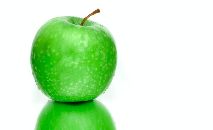 Grüner Apfel mit Wassertropfen auf glänzender Oberfläche vor weißem Hintergrund.
