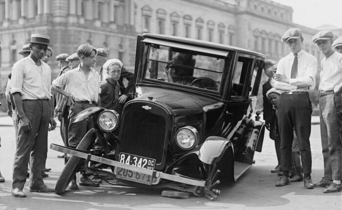 Schwarz-Weiß-Foto von ca. 1920, das eine Gruppe Männer und Jungen zeigt, die ein beschädigtes Auto mit eingeknicktem Vorderrad betrachten.