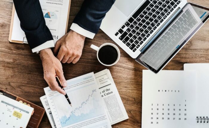 Mann analysiert Finanzdiagramme auf einem Papier, während er an einem Holztisch mit Laptop und Kaffee arbeitet.