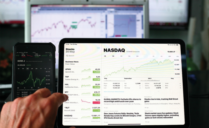Hand zeigt auf Tablet mit NASDAQ-Chart; daneben Smartphone mit Dow Jones und Kursdaten.