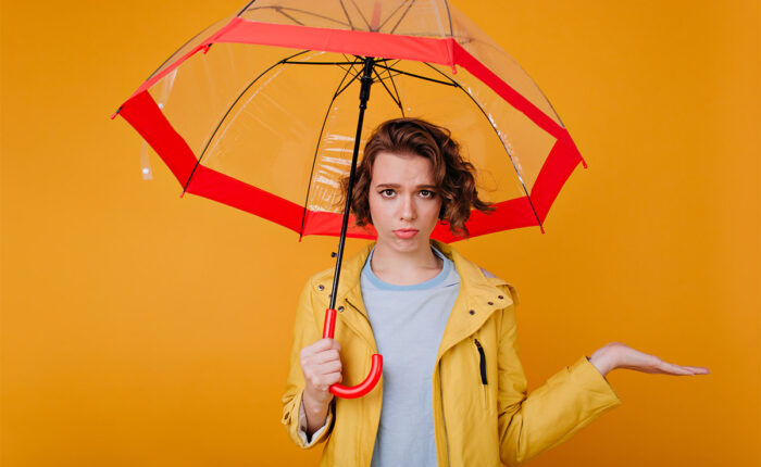 Frau mit gelber Regenjacke und rotem Regenschirm blickt skeptisch, vor orangem Hintergrund.