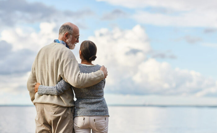Älteres Paar steht umarmt am Wasser und blickt gemeinsam in die Ferne unter einem bewölkten Himmel.