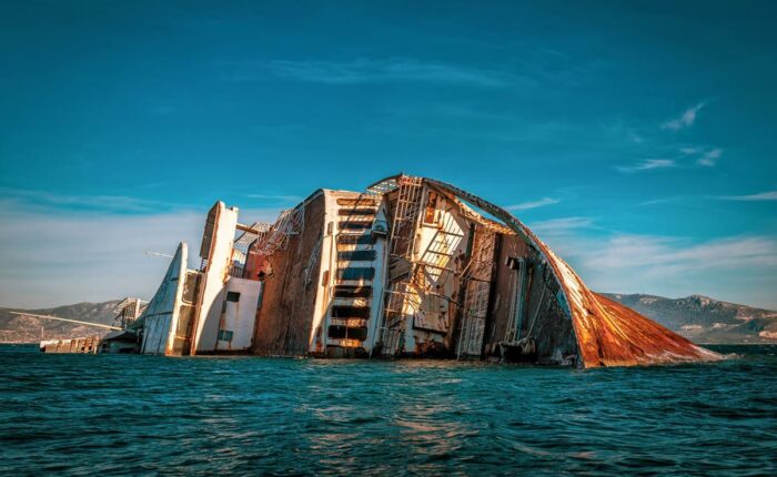 Ein großes, teils untergegangenes Schiff mit rostigen Strukturen ragt aus dem Meer, mit einer Küstenlandschaft im Hintergrund.