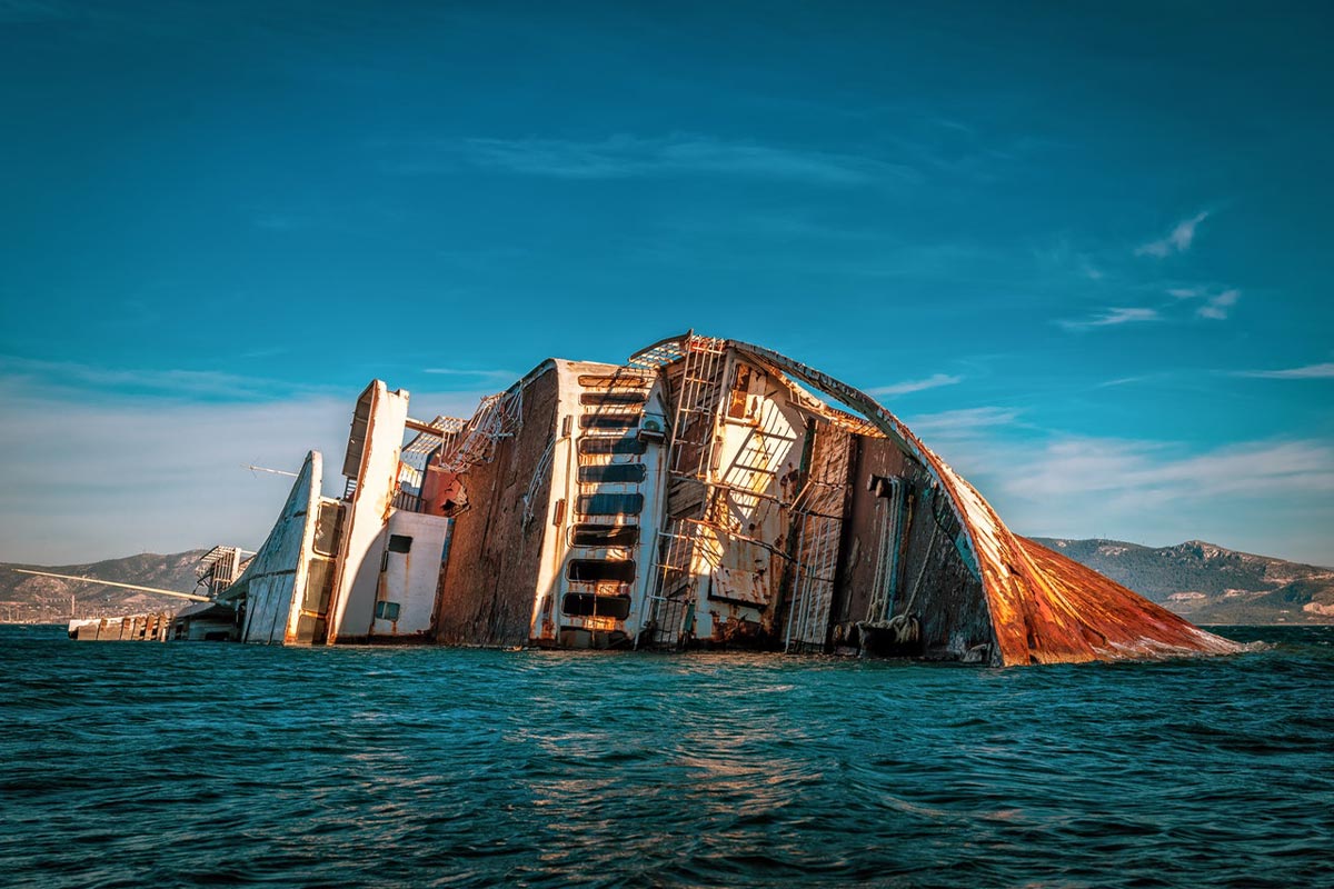 Ein großes, teils untergegangenes Schiff mit rostigen Strukturen ragt aus dem Meer, mit einer Küstenlandschaft im Hintergrund.
