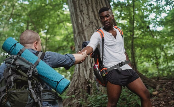 Zwei Wanderer im Wald, einer hilft dem anderen mit ausgestreckter Hand beim Aufstieg, beide mit Rucksäcken und Ausrüstung.